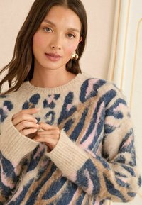 Mīkstas auduma džemperis ar bēša krāsu un abstraktiem zilajiem, rozā un brūnajiem rakstiem. Ribotas aproces un apaļa kakla izgriezums.