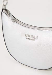 Borsa a tracolla metallica argento con finitura texturizzata, forma curva, chiusura con zip superiore e logo "GUESS" impresso sul davanti.