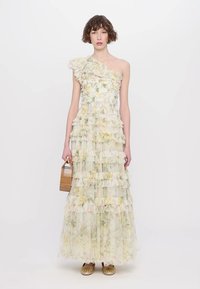 Maxi-jurk van transparant materiaal met bloemenpatronen, gerimpelde lagen, een schouderontwerp en een lichte pastel kleurenpalet. Gouden instapschoenen en een boodschappenmand.