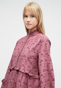 Ροζ μπλούζα από δαντέλα με ψηλό λαιμόκοψη, βολάν στο τελείωμα και περίπλοκα μοτίβα φύλλων. Κατασκευασμένη από υφασμα με υφή και λεπτομερή σχεδιασμό.