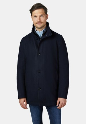Mann trägt einen dunkelblauen Mantel mit Knöpfen über einem hellblauen Reißverschluss-Pullover und Jeans, steht vor einem schlichten hellen Hintergrund.