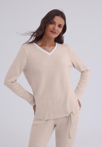 Beżowy sweter w kształcie litery V, wykonany z miękkiego materiału, z długimi rękawami, białym wykończeniem przy dekolcie oraz luźnym krojem z prostym dołem.