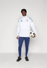 Vit sportjacka med hög krage och blå accenter, kombinerad med marinblå byxor med vita ränder, håller en svartvit fotboll.