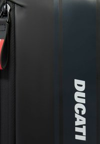 Sac noir avec matériau texturé, large fermeture éclair et logo blanc "DUCATI" bien visible. Présente des rayures sombres discrètes sur le côté.
