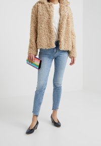 Chaqueta de texturizado de piel de color marrón, blusa blanca, jeans ajustados de color azul claro, tacones negros de punta afilada, sosteniendo un bolso de mano de rayas multicolores.