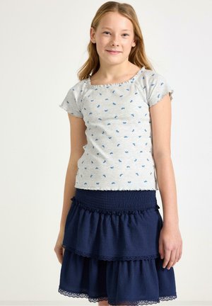 Nuori tyttö, jolla on pitkät vaaleat hiukset, pukeutuneena vaaleanharmaaseen T-paitaan, jossa on sininen kukkakuosi, ja tummansiniseen kerrokselliseen hameeseen, seisoo yksinkertaisen taustan edessä.