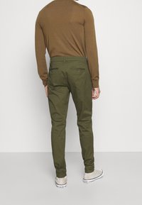 Pantaloni verde oliva in cotone, con vestibilità aderente, tasche posteriori con zip e orli rifiniti. Abbinati a sneaker beige.