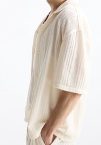 Mužský model nosí krémové krátké síťované košili na knoflíky s bílým tričkem pod ní, s rukou v kapse, na bílém pozadí.