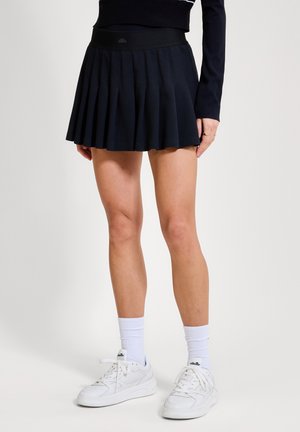 Sieviete, kas valkā melnu plīstošu minisvārku, melnu garu piedurkņu topu, baltas zeķes un baltas sporta kurpes uz vienkārša fona.