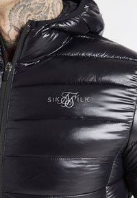 Sort polstret jakke med en skinnende, quiltet tekstur, hætte og en central lynlås. "SikSilk"-logo i hvid på brystet.