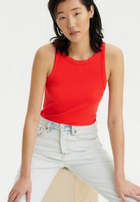 Top sin mangas rojo con textura acanalada y cuello redondo, combinado con jeans de tiro alto azul claro, sentado sobre un bloque de madera de color neutro.