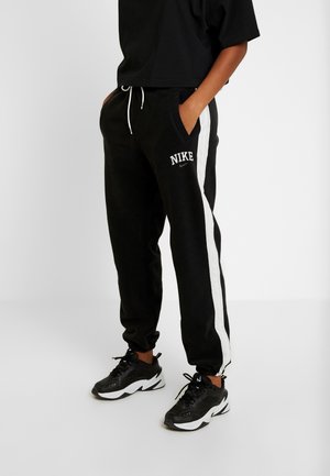 Pantalones deportivos negros con franjas laterales blancas, cinturilla con cordón y bolsillos. Logotipo "NIKE" impreso en el muslo izquierdo. Llevados con zapatillas negras.