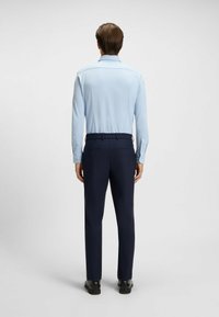 Hellblaues Langarmhemd mit geradem Kragen, kombiniert mit dunkelblauen Hosen mit elastischem Bund und seitlichen Taschen, von hinten betrachtet.