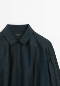 Chemise bleu marine foncé à manches longues, boutonnée, avec col pointu, texture lisse et étiquette de marque Massimo Dutti à l’intérieur du col.