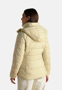 Svetlo béžová pufferek bunda s horizontálnym prešívaním, kapucňou a zipsovým uzatváraním. Obsahuje bočné vrecká a mäkký, textúrovaný povrch.