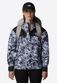 Blue and black patterned jacket with a zip front, black accents, and a high collar, worn with a gray backpack and black pants.