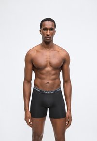 Μυώδης άνδρας όρθιος, στραμμένος προς τα εμπρός, φοράει μαύρα εσώρουχα Calvin Klein boxer briefs, πάνω σε απλό λευκό φόντο.