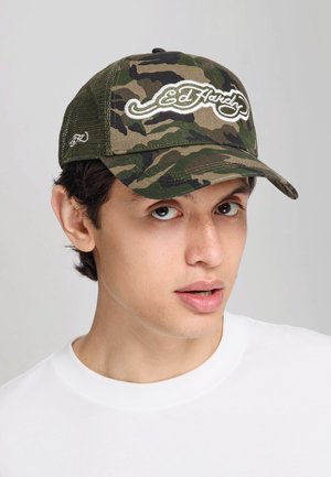 Joven con camisa blanca y gorra de béisbol Ed Hardy verde con camuflaje, mirando ligeramente hacia la derecha sobre un fondo liso y claro.