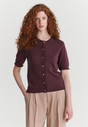 Mujer con el pelo largo, rizado y rojo, que lleva un cárdigan burdeos de manga corta y botones y pantalones beige sobre un fondo liso.