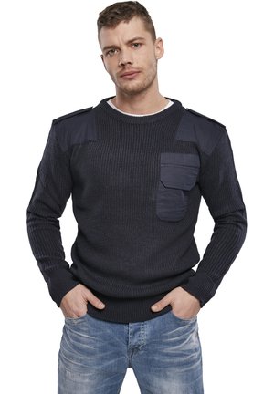 Maglione lavorato a maglia navy con texture a coste, toppe sulle spalle e tasca frontale. Abbinato a jeans in denim chiaro, mani in tasca.