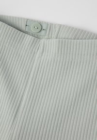 Vihreä ribbikangas, jossa on nappikiinnitys, teksturoitu pinta ja sileä vyötärö. Näkyvät ompeleet korostavat.