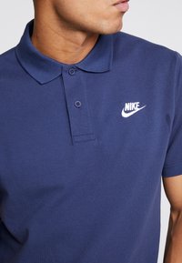 Man som bär en marinblå Nike polo med vitt logotyp, vänd åt sidan mot en enkel bakgrund.