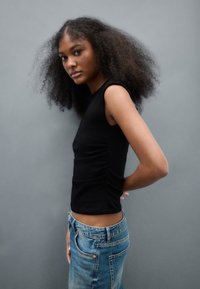 Musta vartalonmyötäinen toppi lyhyillä hihoilla, jossa sivussa rypytykset. Yhdistetty korkeavyötäröisiin vaaleansinisiin farkkuihin. Yksinkertainen harmaa tausta.