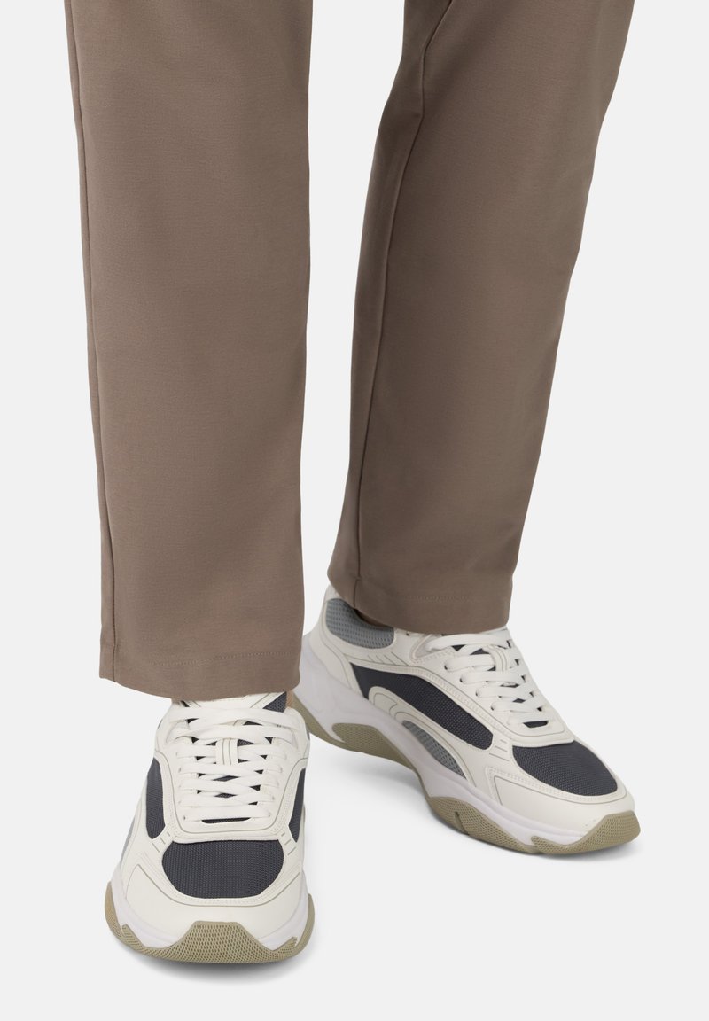 Valged ja hallid spordijalatsid koos võrgusilma aktsentidega, ümar varvas, paksud tallad ja paelad, koos pruunide sirgete pükstega.