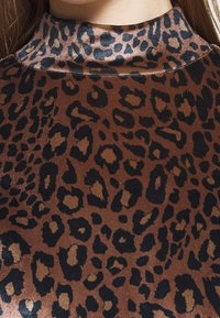 Tessuto marrone con un design a stampa leopardata nera e accenni di beige. Il materiale testurizzato ha una leggera lucentezza, con un alto colletto.