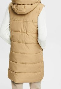 Gilet imbottito beige con un ampio cappuccio e design a righe orizzontali. Presenta una cerniera laterale e maniche bianche texturizzate.