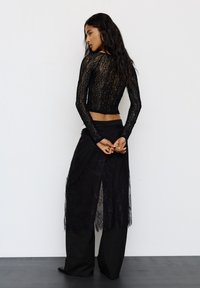 Femeie cu păr lung și închis la culoare, purtând un crop top din dantelă neagră cu mâneci lungi, o fustă din dantelă neagră și pantaloni largi negri, întorcându-se lateral cu mâinile încrucișate la spate.