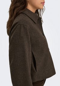 Femme de profil portant une veste texturée marron foncé avec un large col et une poche latérale sur un fond clair.
