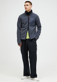 Man standing wearing gray fleece jacket with chest pocket, black pants, neon green shirt, black neck gaiter, and white shoes, looking left.