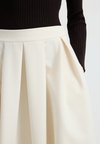 Jupe plissée de couleur crème avec une texture lisse et une taille ajustée. Le tissu est léger et tombe doucement, avec des plis profonds.