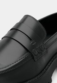Black leather loafer with smooth finish, featuring a decorative strap and stitched accents. Rounded toe and textured upper detailing.