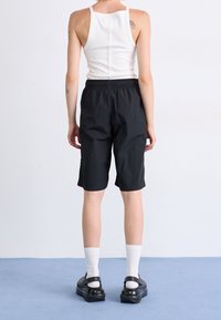 Person steht auf blauem Boden und trägt schwarze kniehohe Shorts, weißes Tanktop, weiße Socken und schwarze Slipper, Ansicht von hinten.