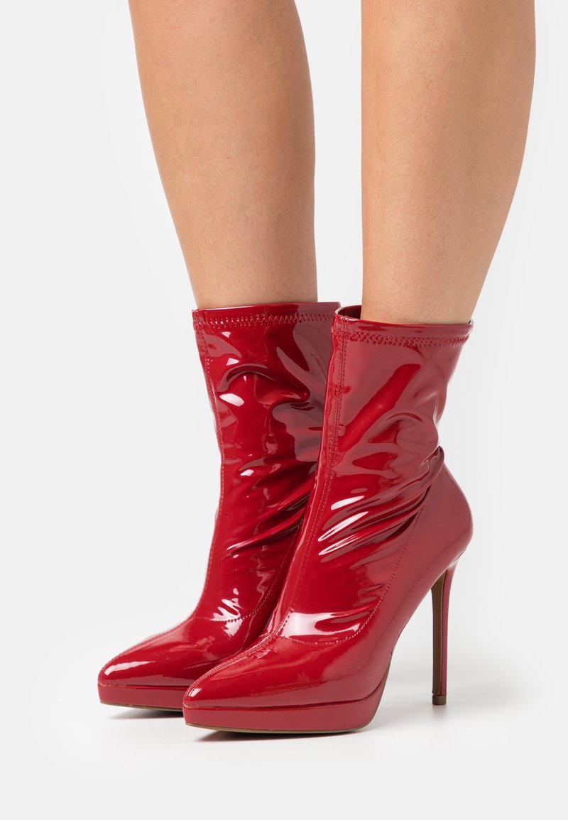 Bottes à cheville en cuir verni rouge brillant avec bouts pointus et talons aiguilles portées sur des jambes nues sur un fond uni.