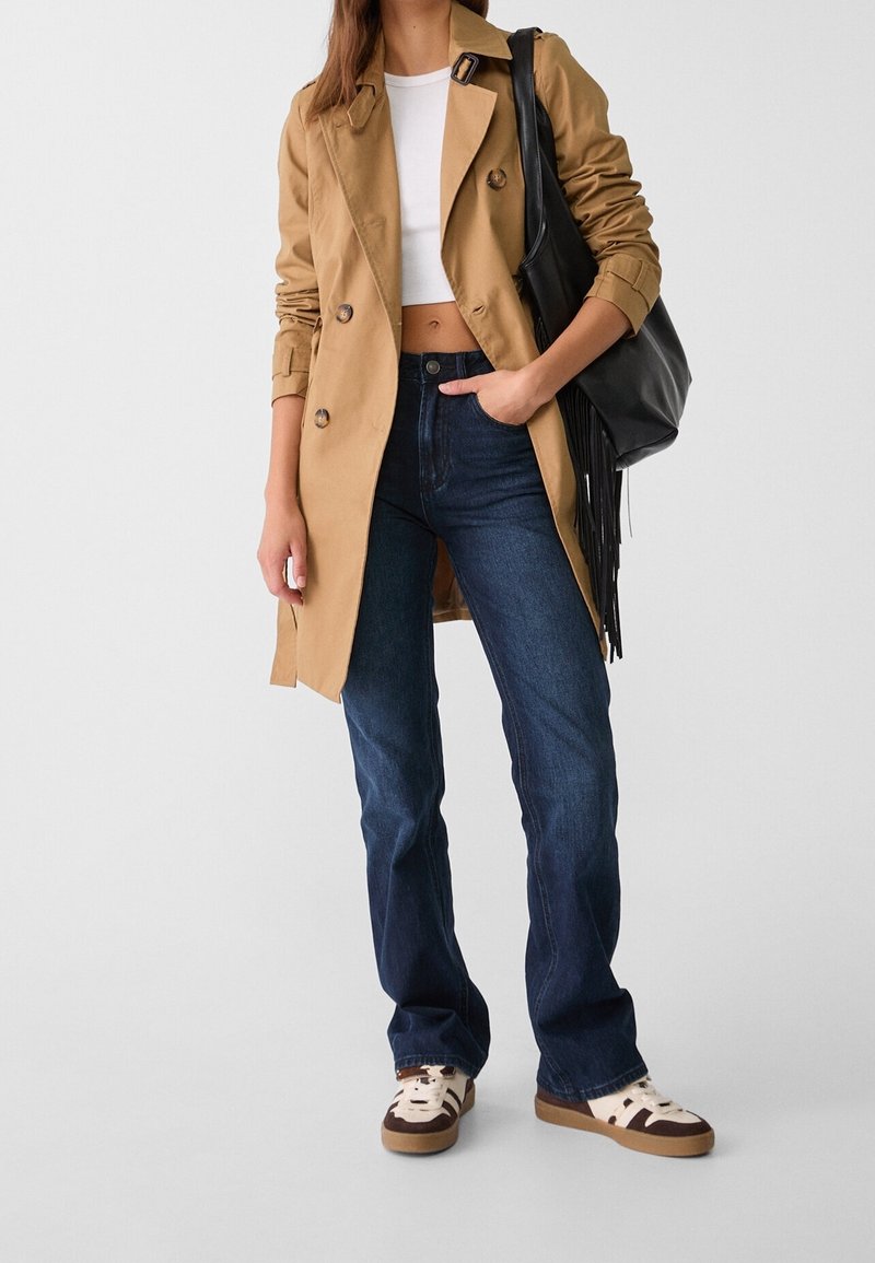 Femme portant un trench beige, un top court blanc, un jean bleu foncé, des baskets, et portant un sac à bandoulière noir à franges.