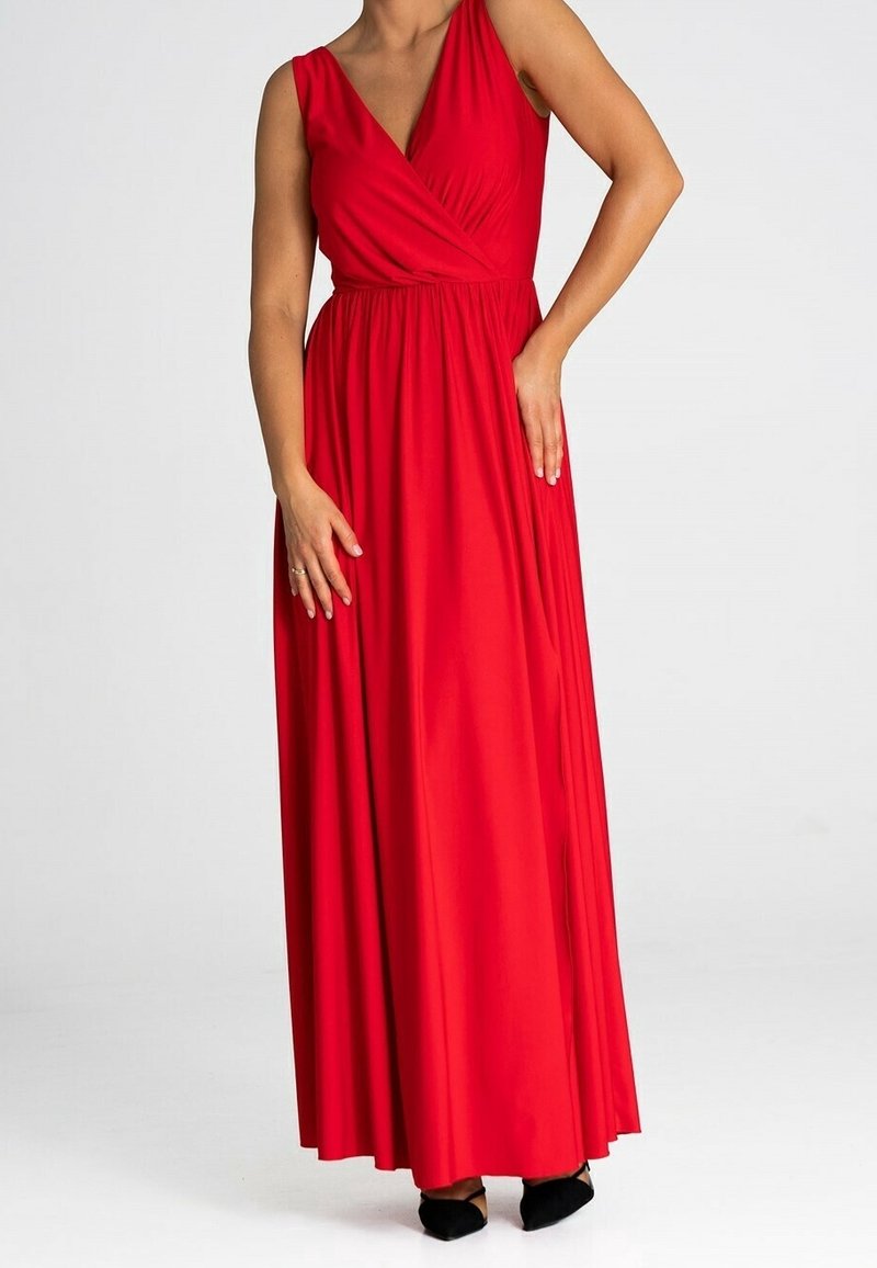 Femme portant une robe maxi rouge sans manches avec un décolleté en V et des chaussures noires pointues, debout devant un fond uni.