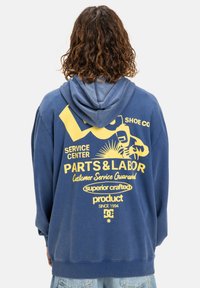 Modrá mikina s kapucňou so zmäkčeným povrchom. Na zadnej strane sú žlté grafické texty a obrázky. Vyrobená z mäkkej tkaniny, s dlhými rukávmi.