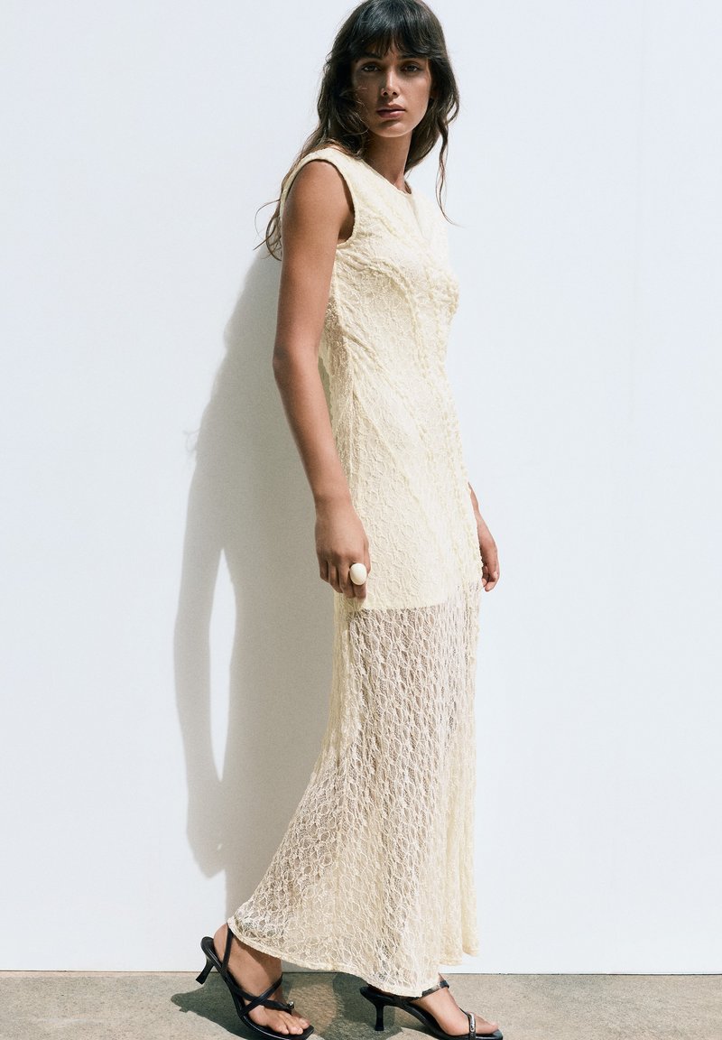 Femme portant une robe maxi en dentelle crème sans manches avec un bas transparent et des sandales à talons noires, debout contre un mur blanc avec une ombre.