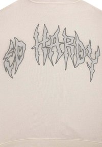 Felpa beige con testo grigio a zig zag "ED HARDY" sulla schiena. Presenta una texture a coste e un design a girocollo. Stile minimalista.