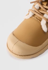 Zapato impermeable color marrón claro con una puntera de goma, parte superior de cuero texturizado y acentos de lana beige. Presenta cordones beige y una suela con patrón.