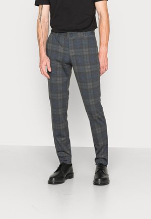 Homme portant un pantalon slim à carreaux gris et bleu, des chaussures noires et une chemise noire, debout devant un fond clair uni.