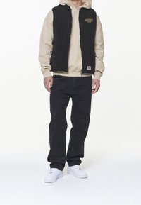 Czarna kamizelka z polaru na beżowej bluzie z kapturem, w zestawieniu z ciemnymi jeansami i białymi butami sportowymi. Kamizelka ma zamek błyskawiczny oraz detal z logo.