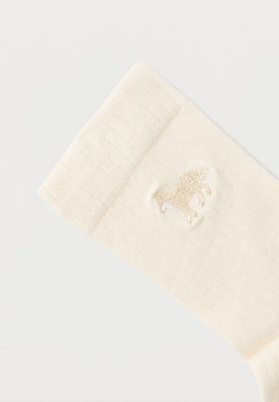 Κρέμ κάλτσα με ριμπ επένδυση και κεντημένο μπεζ μοτίβο δεινοσαύρου κοντά στο πάνω μέρος.