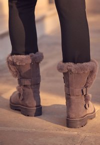 Bottes en daim gris avec bordure en fourrure, sangles et boucles, dotées d'une semelle épaisse. Le design est court et arrondi à l'avant.