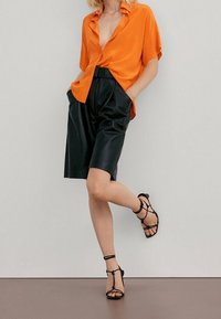 Orange short-sleeve blouse with a knotted hem paired with black leather shorts and strappy black sandals, standing against a neutral background.