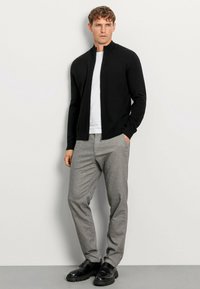 Schwarzer Reißverschluss-Pullover über einem weißen Hemd, kombiniert mit grauen Hosen und schwarzen Schuhen. Der Stoff ist glatt und hat eine figurbetonte Passform.