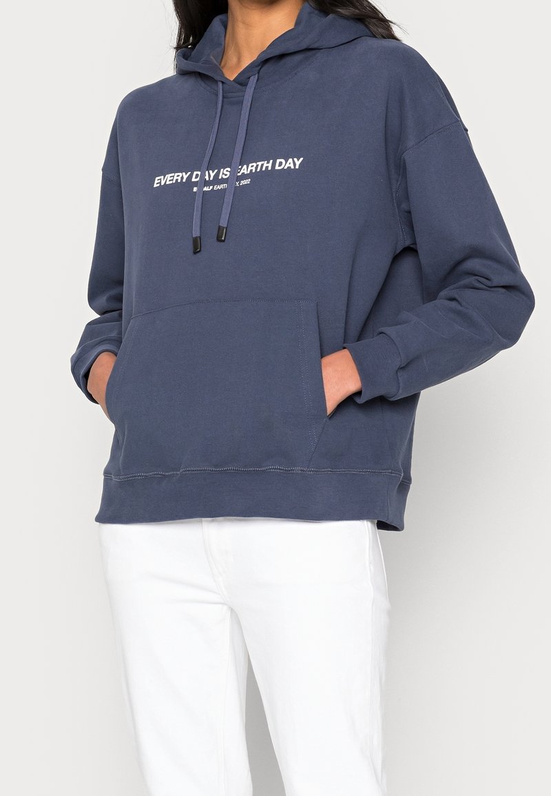 Mörkblå hoodie i mjukt tyg, med en ficka framtill, snörning och vit text som säger "VARJE DAG ÄR JORDENS DAG."